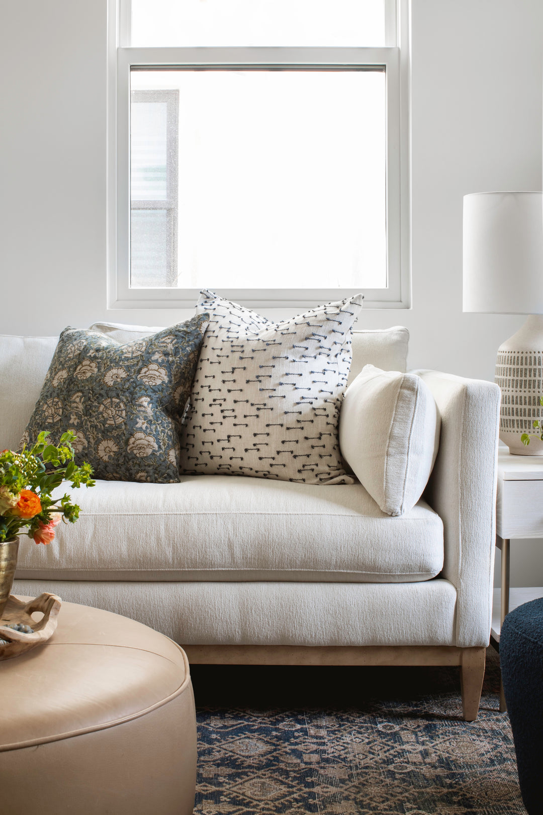 Modern white sofa with patterned pillows, vintage area rug, and florals on round ottoman in a bright living room.