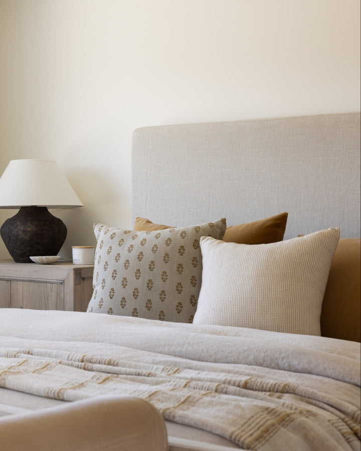 Bedroom with a bed featuring patterned and solid pillows, a lamp on a nightstand, and neutral walls.